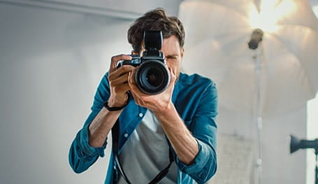 A young male professional photographer holds his DSLR camera to take a shot inside a photography studio with backlighting